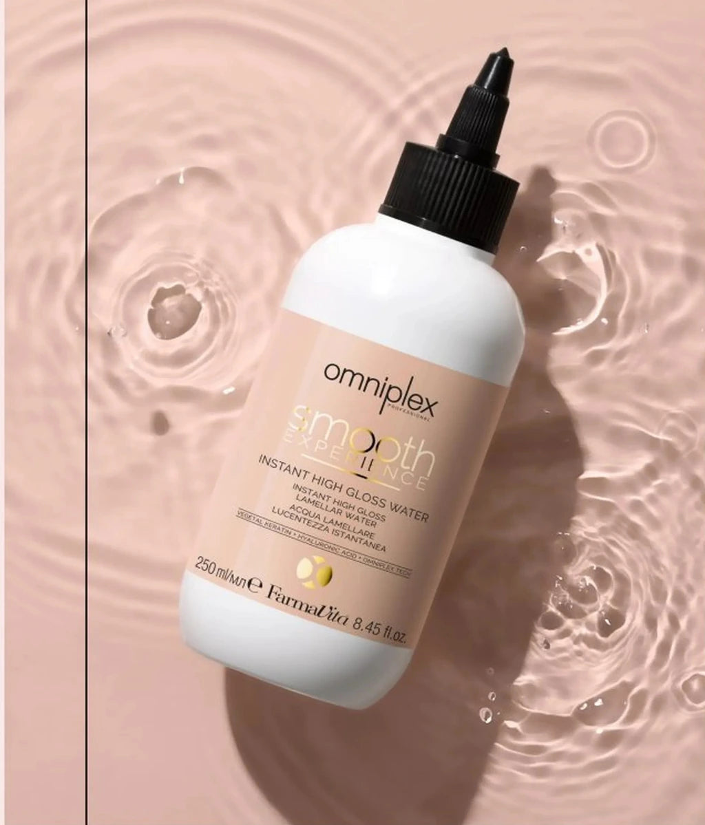 A white bottle of omniplex smooth shine instant high glow water hair serum with a black dropper cap, set against a background of water ripples on a dusty pink surface.